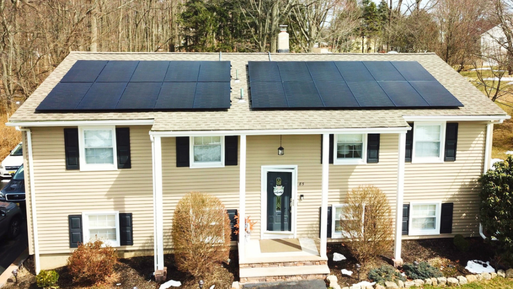 Front view of a beige two-story house with black window shutters and a black front door, featuring two large arrays of black solar panels installed on the roof. The yard is landscaped with bushes and patches of snow, and trees are visible in the background.